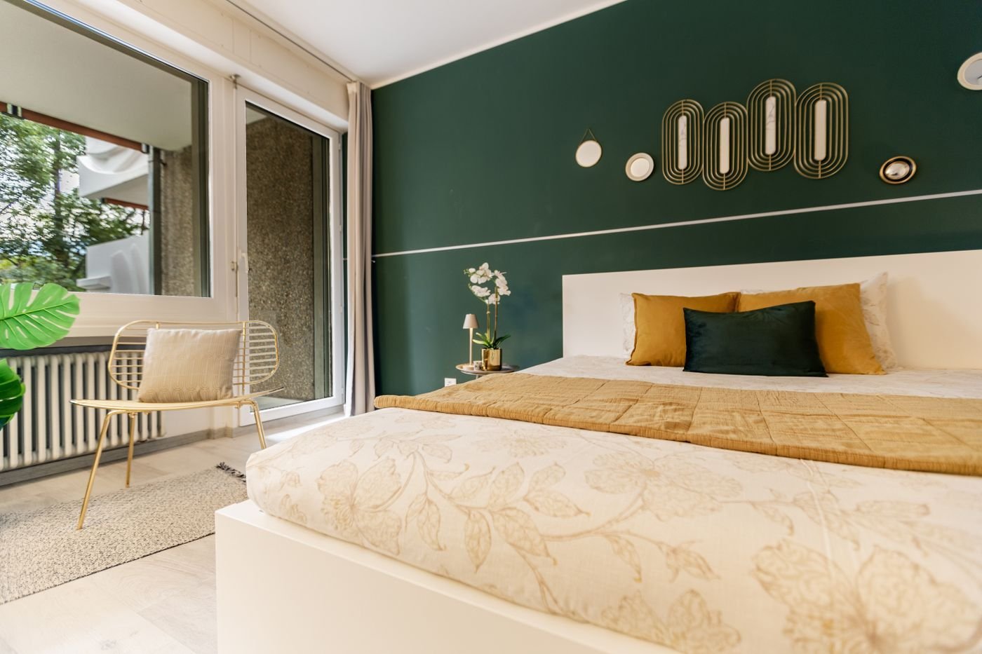 A bright bedroom with a deep green feature wall, pale bedding and warm gold accents